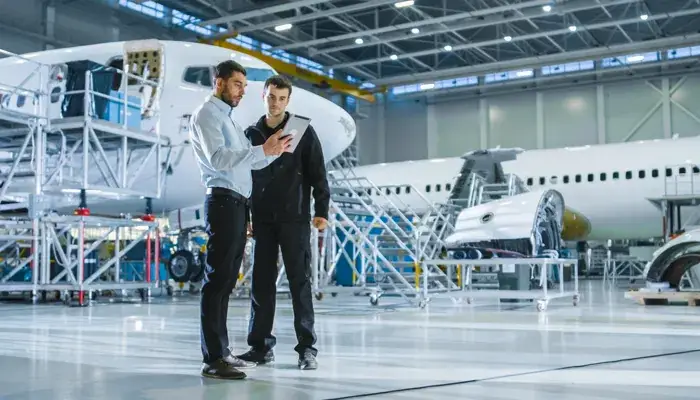 Two engineers in an aerospace manufacturing facility discussing quality control measures in front of a partially assembled aircraft.