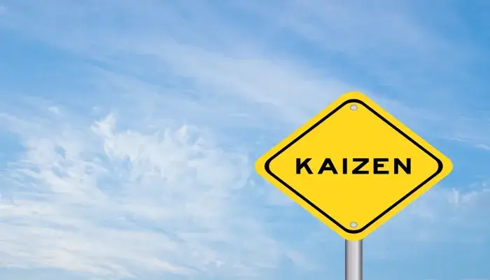 A yellow road sign with the word 'Kaizen' against a bright blue sky with light clouds.