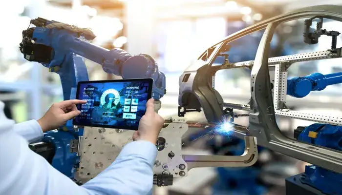 An engineer uses a tablet to monitor and control robotic arms assembling a car frame in a smart factory.