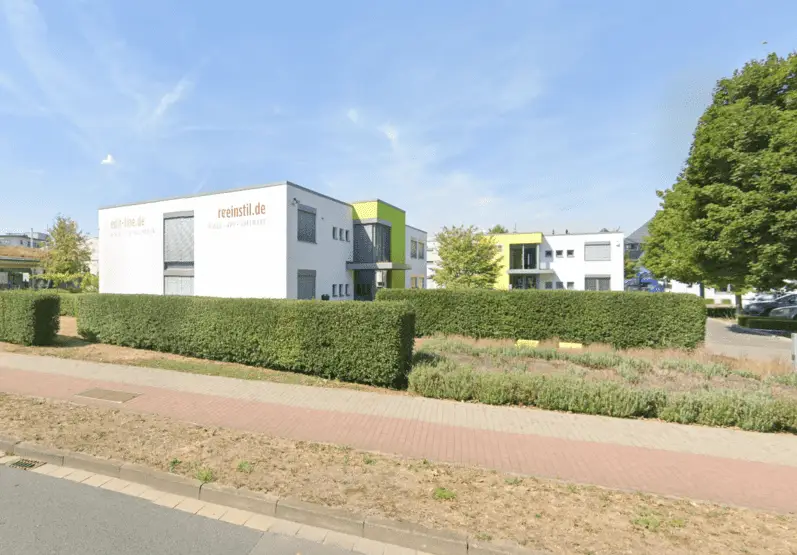 The image shows a modern office building with "reinstil.de" signage, surrounded by trimmed hedges and greenery, on a sunny day with a clear sky.