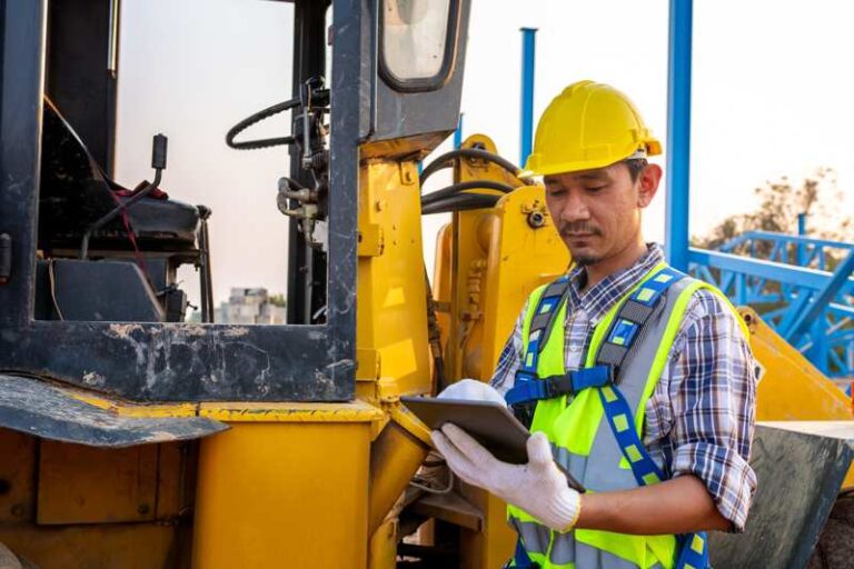 A construction worker wearing a yellow helmet holding a tablet in front of a yellow machine.