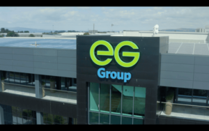 The image shows the exterior of a modern corporate building with the logo "EG Group" prominently displayed. The building features large glass windows, a balcony area, and a rooftop covered in solar panels, suggesting a focus on sustainability.