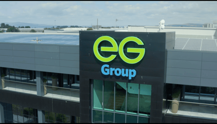 The image shows the exterior of a modern corporate building with the logo "EG Group" prominently displayed. The building features large glass windows, a balcony area, and a rooftop covered in solar panels, suggesting a focus on sustainability.