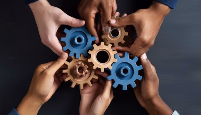 Diverse hands connecting blue and bronze gearwheels, symbolizing collaboration and alignment in standard work processes.
