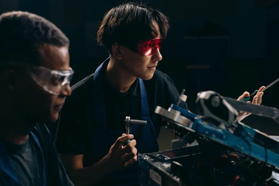Two young people working with tools in a workshop, one wearing goggles and focused on their task.