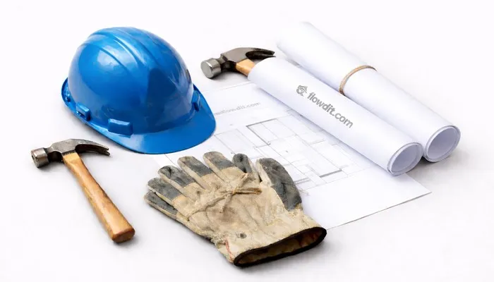 Construction tools on a light grey surface showing maintenance standards with a blue hard hat, worn work gloves, a hammer, and rolled technical drawings labeled flowdit.com